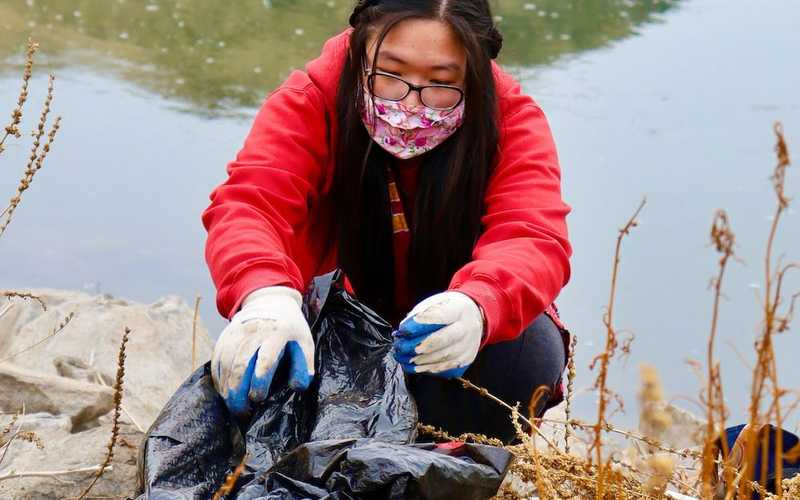 a woman in a red coat, gloves and mask placing recyclables into a black bag