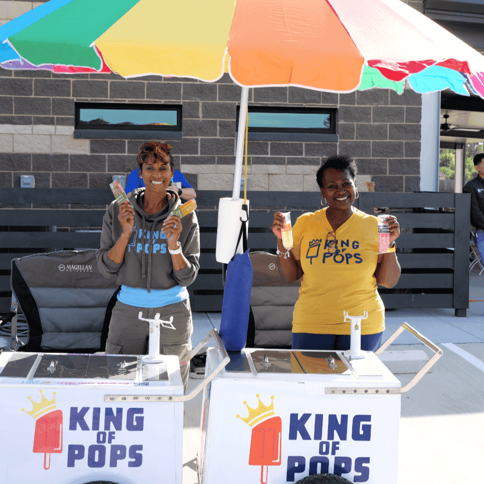 Two smiling women stand behind King of Pops carts under a rainbow umbrella, each holding up two packaged popsicles while posing for a photo.