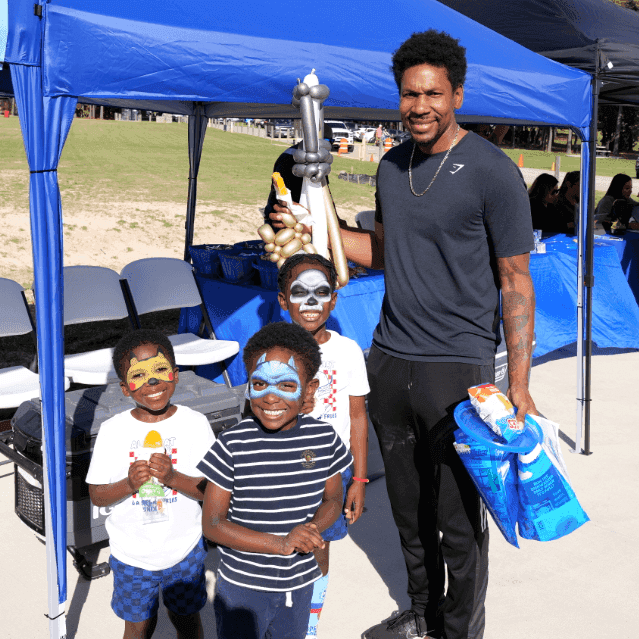 A man stands with three young boys featuring superhero face paint. One child holds a balloon sculpture while they pose together in front of event tents.